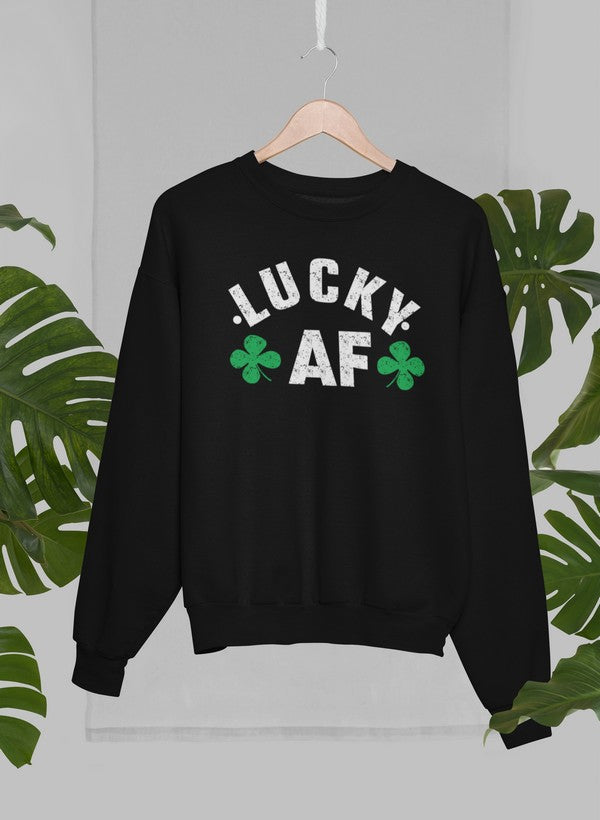 Black crewneck sweatshirt with white lettering that says “LUCKY AF” with 2 four-leaf clovers handing on a wooden hanger with green leaves in the background.