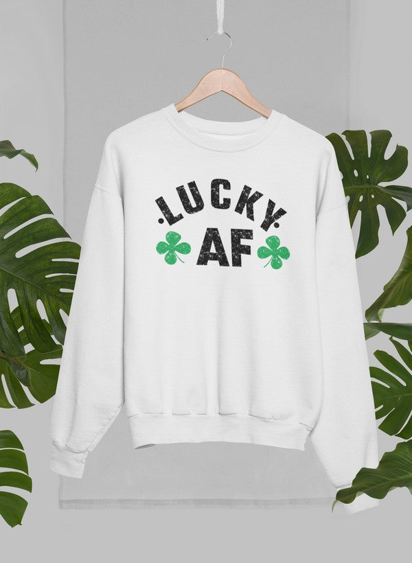 White crewneck sweatshirt with black lettering that says “LUCKY AF” with 2 four-leaf clovers handing on a wooden hanger with green leaves in the background.