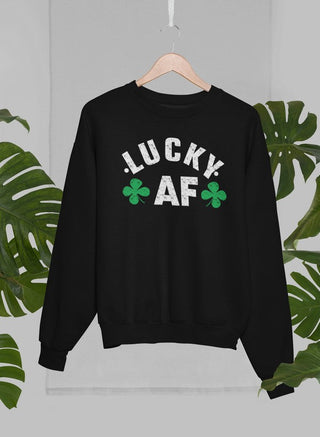 Black crewneck sweatshirt with white lettering that says “LUCKY AF” with 2 four-leaf clovers handing on a wooden hanger with green leaves in the background. 