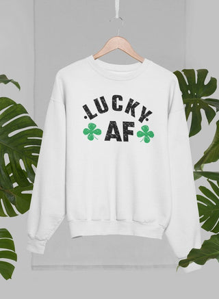 White crewneck sweatshirt with black lettering that says “LUCKY AF” with 2 four-leaf clovers handing on a wooden hanger with green leaves in the background. 