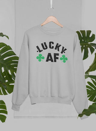 Grey crewneck sweatshirt with white lettering that says “LUCKY AF” with 2 four-leaf clovers handing on a wooden hanger with green leaves in the background. 